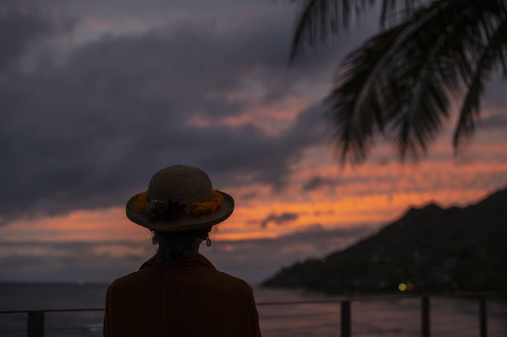 A bright orange and light purple sunset dips behind Leahi (Diamond Head) over a calm ocean. IN the foreground is a palm tree to the right of the picture. A woman in a hat stands in silhouette in the foreground looking at the sunset.