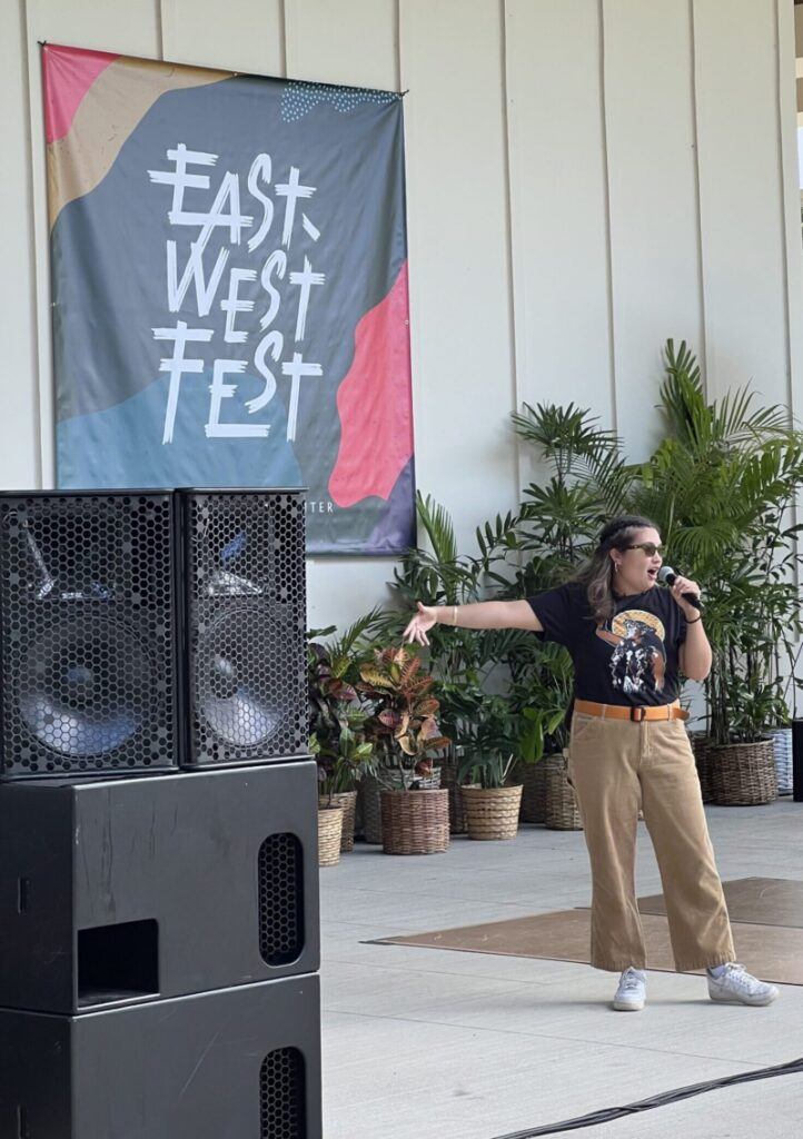 Performing artist Inalihi stands holding a microphone performing their spoken word music. To the left of the photo is a large black speaker and in the background is a multi-colored banner with the words, "East-West Fest" and potted tropical plants below the banner. 