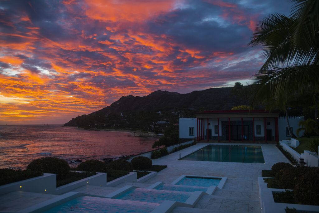 A multi-colored sky of bright yellow, orange, pink and purple sets behind Leahi (Diamond Head) reflecting bright colors on the calm ocean. In the right hand-side of the photo is the Shangri La plahyouse during sunset  and the cascades of blue water leading to the pool and playhouse. 
