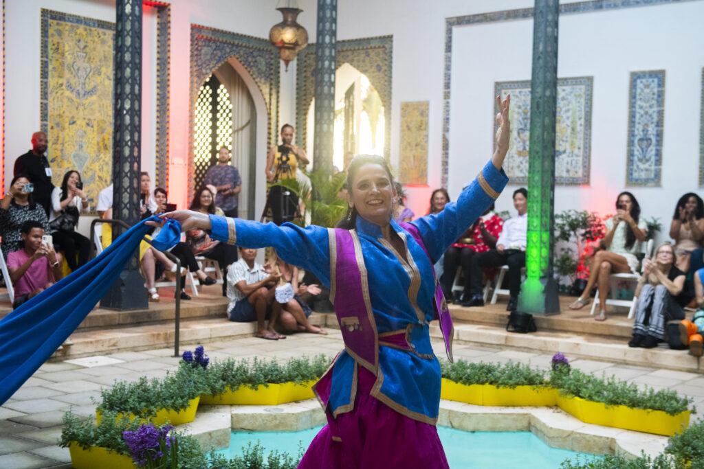 A woman dressed in a blue, magenta, and gold outfit dances holding a blue scarf trailing behind her. The dancer is in front of a star-shaped fountain surrounded by yellow planters with plants and purple hyacinth flowers. In the background of the photo sits a crowd watching the dancer. 