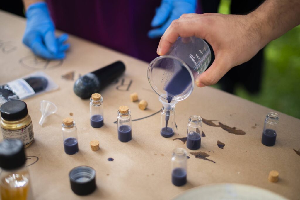 A hand holding a glass beaker pours grey-purple colored ink into small glass bottles with cork stoppers. The bottles are on a brown paper covered table. 