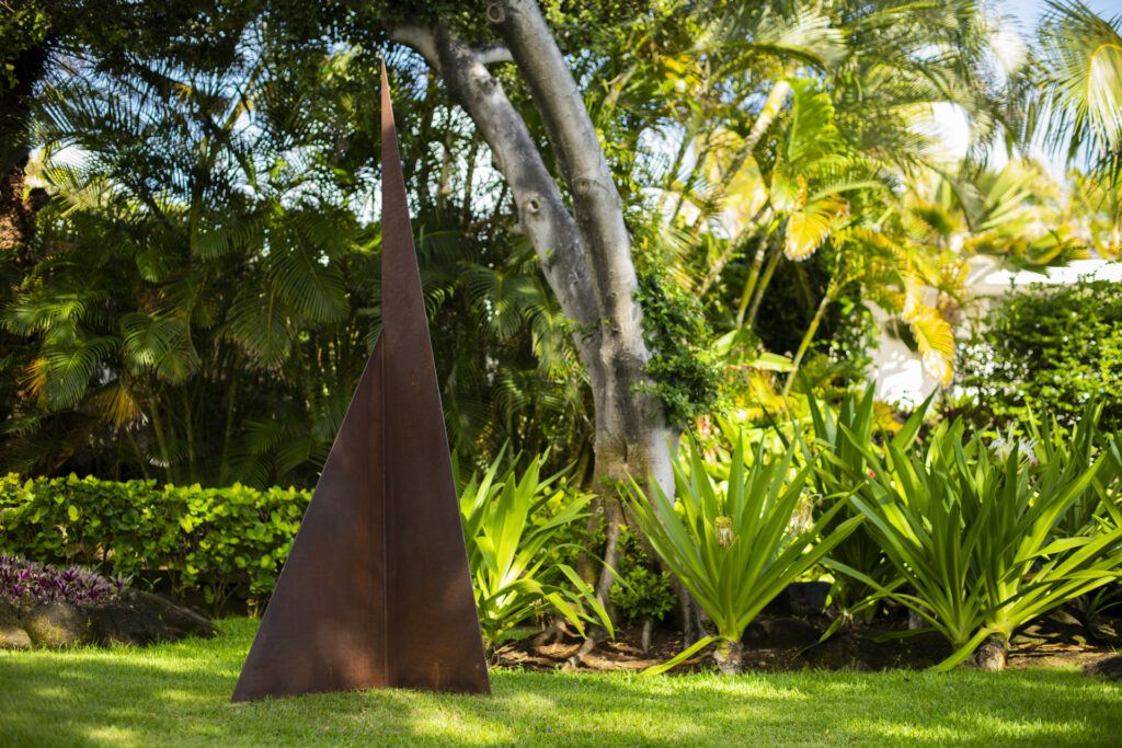 An abstract rusted steel sculpture in a triangular shape stands in the middle of a garden setting at Shangri La